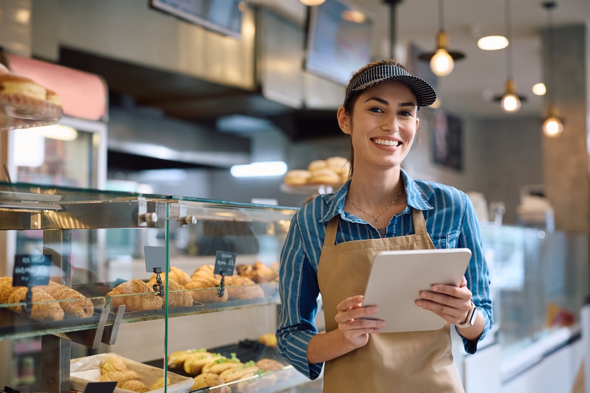 Dueña de una panadería sonriendo con confianza, clientes satisfechos al fondo, ambiente cálido y profesional